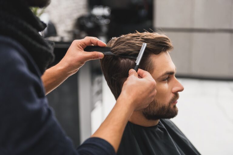 Main tenant un peigne et un rasoir, coupant avec précision les cheveux d'un homme barbu portant une cape de barbier noire.