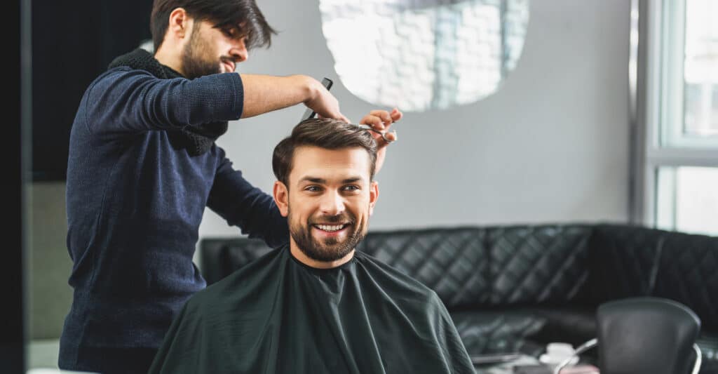 Barbier coupant les cheveux d'un homme souriant et barbu, assis dans un fauteuil de salon de coiffure moderne.