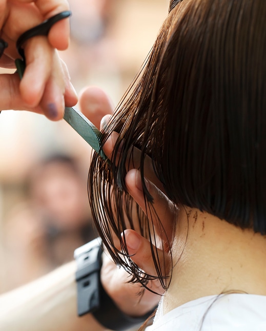 Mains coupant des cheveux noirs mouillés au carré avec des ciseaux et un peigne vert, vue arrière en salon.