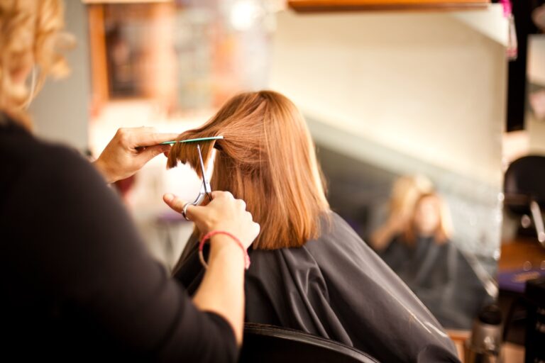 Coiffeuse coupant les cheveux roux d'une cliente avec peigne et ciseaux, vue de dos dans un salon. Reflet flou au miroir.