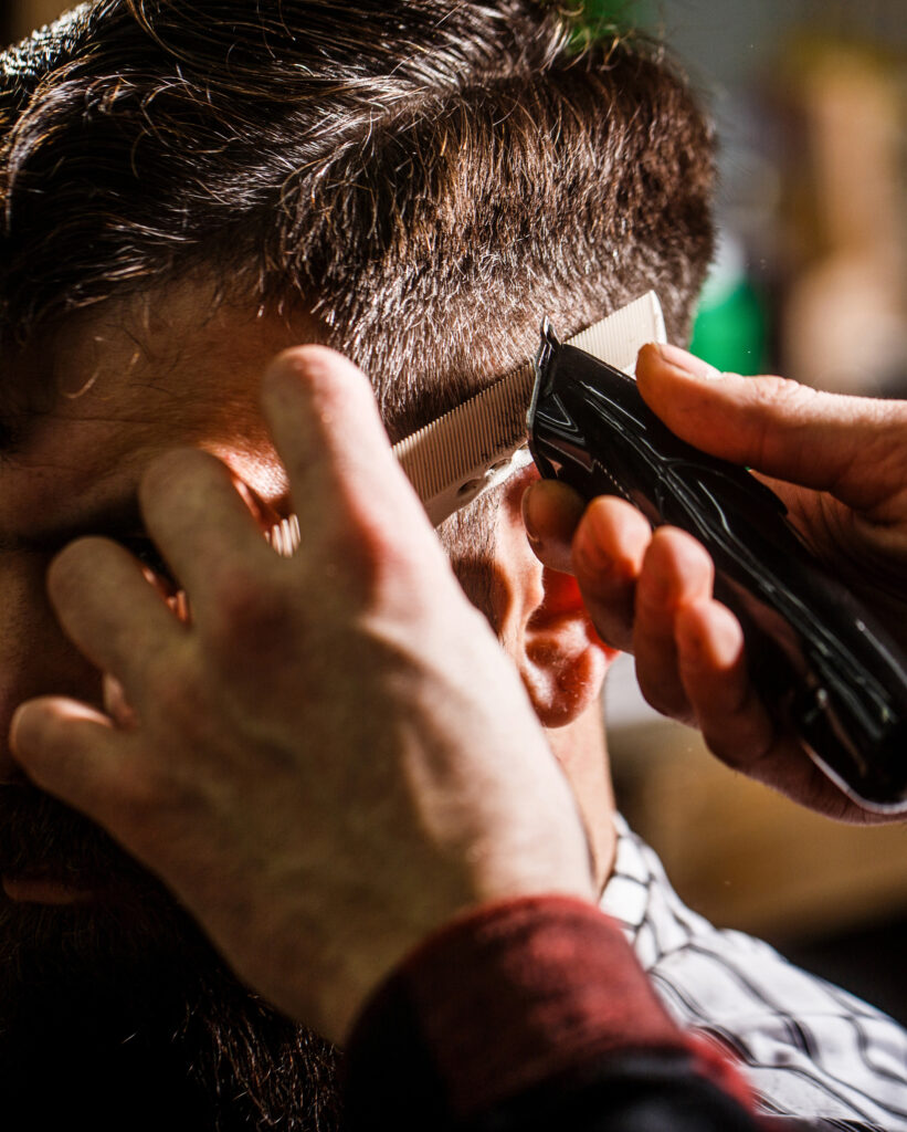 Barbier coupant les cheveux d'un homme aux cheveux foncés avec une tondeuse électrique et un peigne. Gros plan sur l'action.